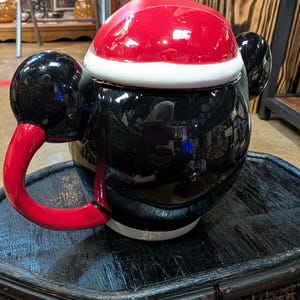 Puede incluir: Una tetera de cer&aacute;mica con forma de personaje, cuerpo negro brillante, asa roja y tapa dise&ntilde;ada como un gorro de Pap&aacute; Noel rojo con borde blanco. La tetera presenta dos orejas circulares negras en los laterales y un acabado reflectante.