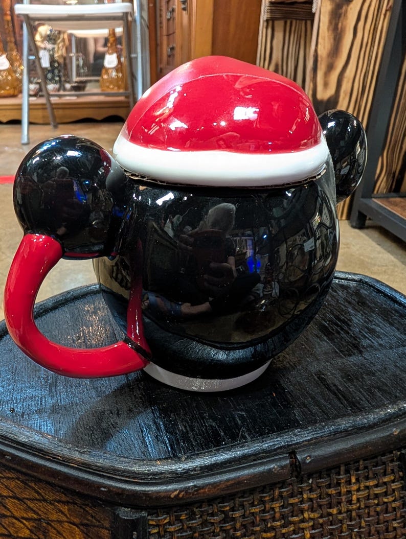 Puede incluir: Una tetera de cer&aacute;mica con forma de personaje, cuerpo negro brillante, asa roja y tapa dise&ntilde;ada como un gorro de Pap&aacute; Noel rojo con borde blanco. La tetera presenta dos orejas circulares negras en los laterales y un acabado reflectante.