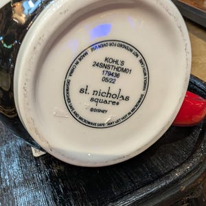 Puede incluir: La parte inferior de una taza de cer&aacute;mica blanca con texto negro que dice KOHL'S 24SNSTHDM01 179436 05/22, st. nicholas square y DISNEY. El texto tambi&eacute;n incluye instrucciones que indican que la taza es apta para lavavajillas y microondas.