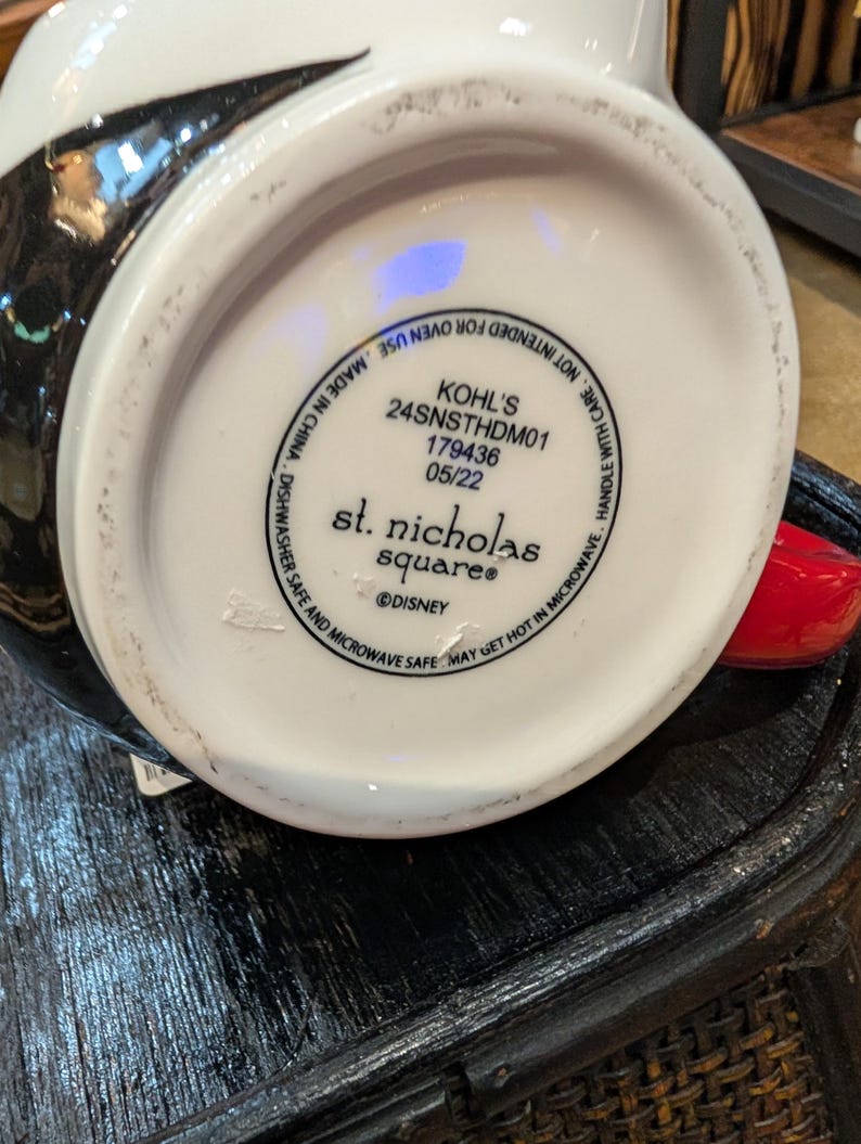 Puede incluir: La parte inferior de una taza de cer&aacute;mica blanca con texto negro que dice KOHL'S 24SNSTHDM01 179436 05/22, st. nicholas square y DISNEY. El texto tambi&eacute;n incluye instrucciones que indican que la taza es apta para lavavajillas y microondas.