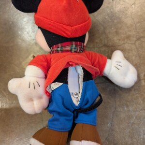 May include: A Mickey Mouse plush toy wearing a red knit hat with a white pom-pom, a red jacket, blue pants, and brown shoes. The plush has black ears, white gloves, and a plaid scarf. The back of the plush has a zipper.