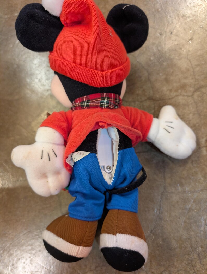 May include: A Mickey Mouse plush toy wearing a red knit hat with a white pom-pom, a red jacket, blue pants, and brown shoes. The plush has black ears, white gloves, and a plaid scarf. The back of the plush has a zipper.