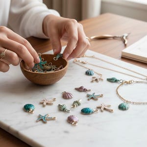 May include: A collection of sea-themed jewelry charms and a bracelet with gold chains and colorful enamel accents. The charms include starfish, seashells, and other marine life designs. The jewelry is displayed on a white marble surface.