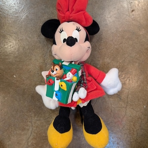 K&ouml;nnte beinhalten: Eine Minnie Mouse Pl&uuml;schpuppe mit rotem Kleid, Schleife und gelben Schuhen. Die Puppe h&auml;lt eine gr&uuml;n-wei&szlig;e Spielzeugkiste mit einer Chip und Chap Figur darin. Die Puppe hat schwarze Ohren und ein l&auml;chelndes Gesicht.