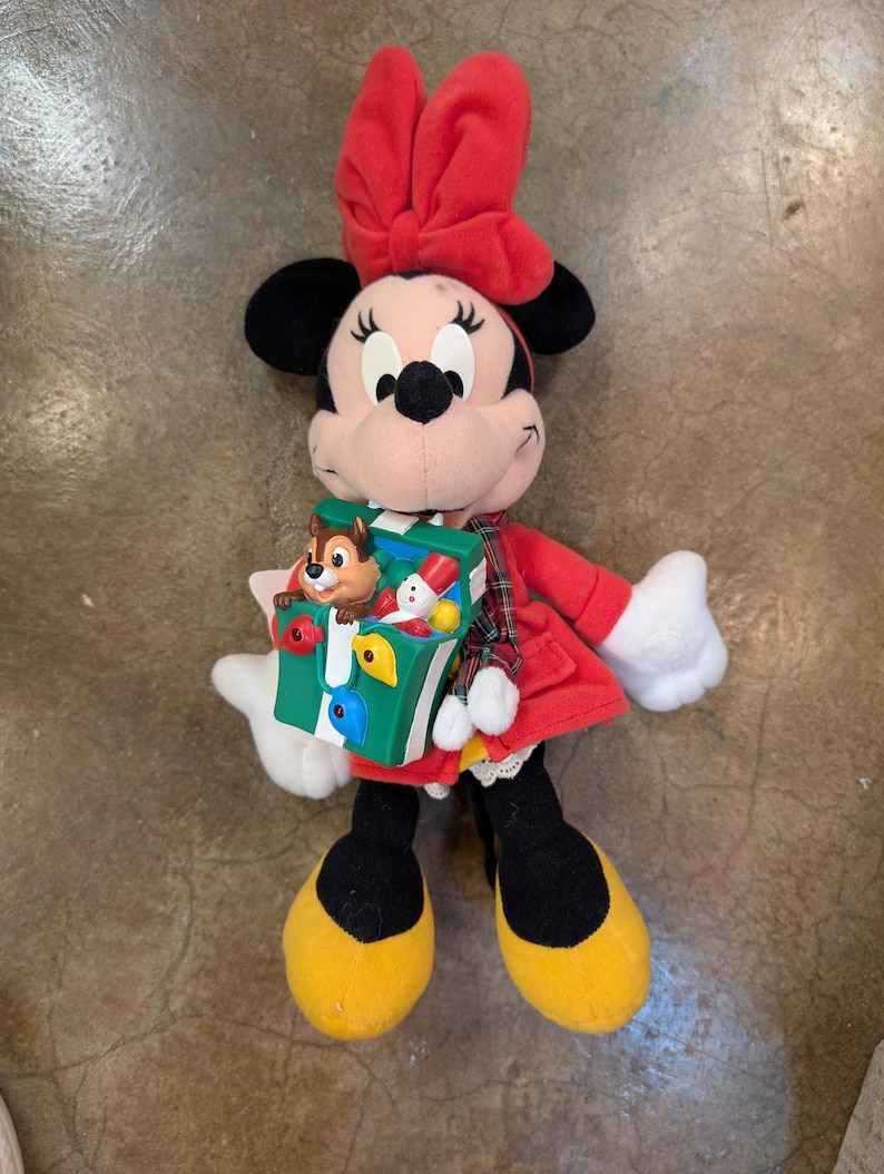K&ouml;nnte beinhalten: Eine Minnie Mouse Pl&uuml;schpuppe mit rotem Kleid, Schleife und gelben Schuhen. Die Puppe h&auml;lt eine gr&uuml;n-wei&szlig;e Spielzeugkiste mit einer Chip und Chap Figur darin. Die Puppe hat schwarze Ohren und ein l&auml;chelndes Gesicht.