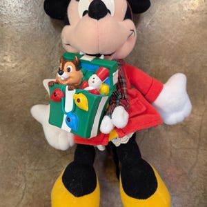 K&ouml;nnte beinhalten: Eine Minnie-Maus-Pl&uuml;schpuppe mit rotem Kleid, Schleife und Schuhen. Die Puppe h&auml;lt eine gr&uuml;ne Spielzeugkiste mit einer Chip-und-Chap-Figur und anderen kleinen Spielzeugen. Die Puppe hat schwarze Ohren und ein rosa Gesicht.
