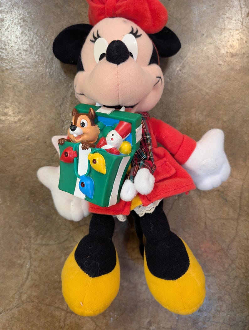K&ouml;nnte beinhalten: Eine Minnie-Maus-Pl&uuml;schpuppe mit rotem Kleid, Schleife und Schuhen. Die Puppe h&auml;lt eine gr&uuml;ne Spielzeugkiste mit einer Chip-und-Chap-Figur und anderen kleinen Spielzeugen. Die Puppe hat schwarze Ohren und ein rosa Gesicht.