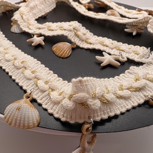 Peut inclure: Garniture tressée crème avec des accents dorés et des coquillages et étoiles de mer suspendus. La garniture est disposée en motif ondulé, avec les coquillages et les étoiles de mer ajoutant un thème côtier.