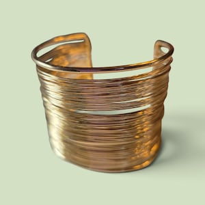 May include: A wide cuff bracelet constructed from multiple thin, metallic gold-toned wires wrapped in a continuous, layered pattern. The design features an open back with a rigid, circular structure and a polished, reflective surface finish.