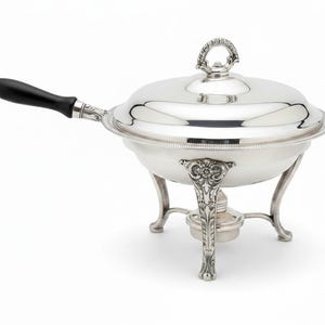 May include: A silver-plated chafing dish featuring a polished round bowl, a matching lid with a decorative ring handle, and three ornate legs with floral detailing. The dish includes a black wooden handle and a central burner underneath the bowl.