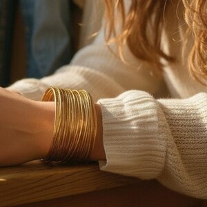 May include: A stack of numerous thin, polished gold-tone metal bangles worn together on the wrist. The bracelets feature a smooth, reflective surface and a uniform circular shape, creating a wide, continuous band of gold around the arm.
