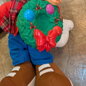 May include: A plush figure of Chip, holding a green Christmas wreath with a red bow and colorful ornaments. Chip is wearing a red shirt, blue pants, and brown shoes with white stripes. The figure has a plaid scarf.