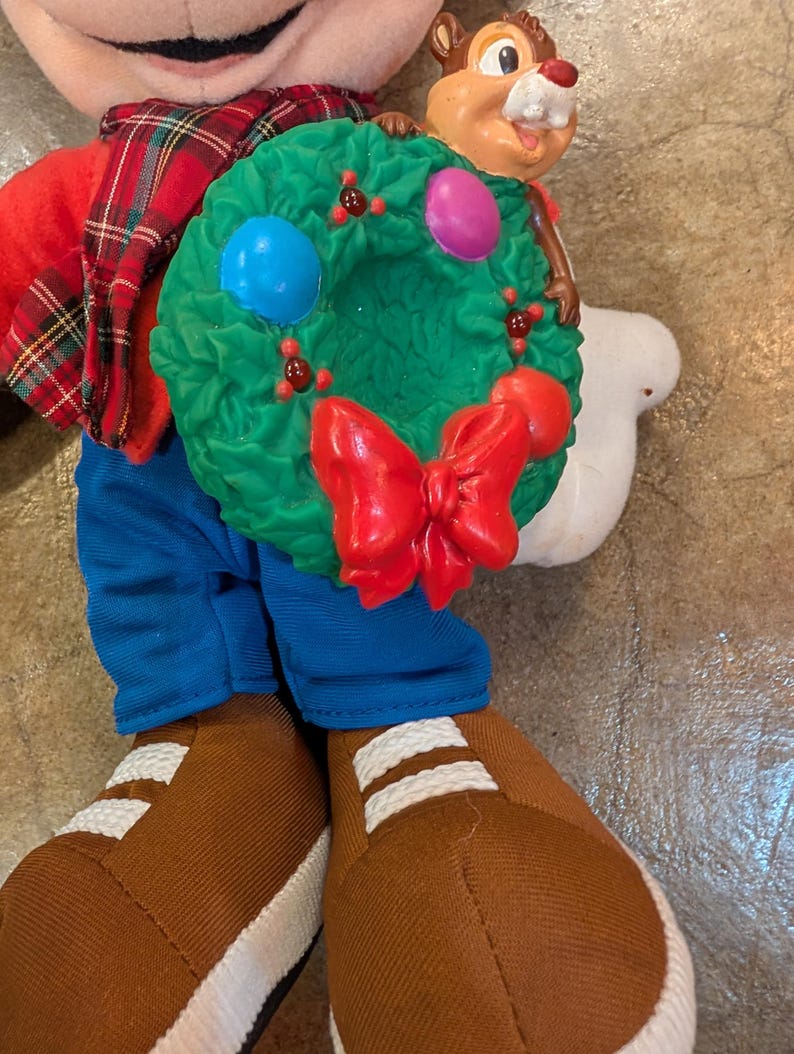 May include: A plush figure of Chip, holding a green Christmas wreath with a red bow and colorful ornaments. Chip is wearing a red shirt, blue pants, and brown shoes with white stripes. The figure has a plaid scarf.