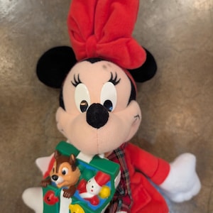 K&ouml;nnte beinhalten: Ein Minnie Mouse Pl&uuml;schtier mit roter Schleife, rotem Mantel und einem gr&uuml;n-wei&szlig;en Spielzeug. Das Spielzeug zeigt Chip und Chap, einen Schneemann und bunte Formen. Das Pl&uuml;schtier hat schwarze Ohren und wei&szlig;e Handschuhe.