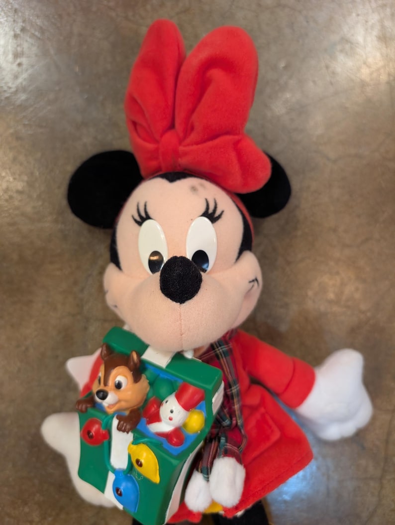 K&ouml;nnte beinhalten: Ein Minnie Mouse Pl&uuml;schtier mit roter Schleife, rotem Mantel und einem gr&uuml;n-wei&szlig;en Spielzeug. Das Spielzeug zeigt Chip und Chap, einen Schneemann und bunte Formen. Das Pl&uuml;schtier hat schwarze Ohren und wei&szlig;e Handschuhe.