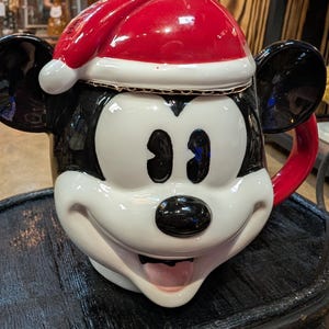 Puede incluir: Una taza de cer&aacute;mica con forma de cabeza de rat&oacute;n de dibujos animados, con un acabado brillante en blanco y negro, grandes orejas redondas y una tapa de gorro de Pap&aacute; Noel rojo. El personaje tiene una expresi&oacute;n amplia y sonriente.