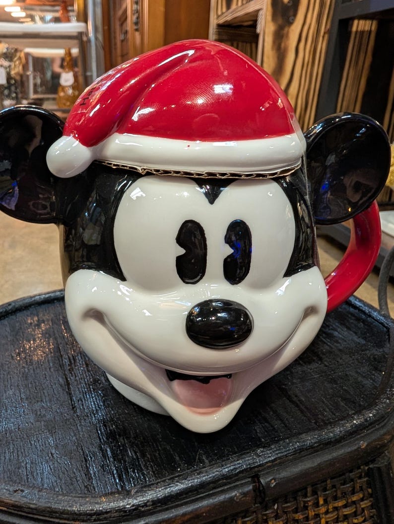 Puede incluir: Una taza de cer&aacute;mica con forma de cabeza de rat&oacute;n de dibujos animados, con un acabado brillante en blanco y negro, grandes orejas redondas y una tapa de gorro de Pap&aacute; Noel rojo. El personaje tiene una expresi&oacute;n amplia y sonriente.