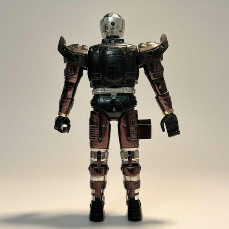 Może przedstawiać: Zabytkowa figurka robota z srebrnym hełmem i akcentami. Robot ma czarny tuł&oacute;w, brązowe ramiona i nogi oraz srebrne detale. Figurka stoi wyprostowana.
