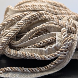 May include: A close-up shot of a coiled trim with a twisted rope design. The trim has a beige fabric base with a twisted rope in shades of beige and cream. This trim is suitable for edging or decorative use in sewing or upholstery projects.