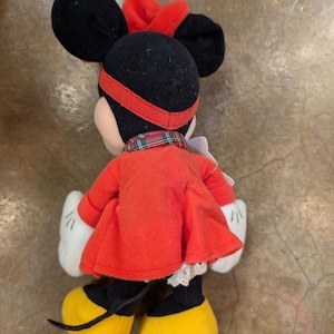 K&ouml;nnte beinhalten: Eine Pl&uuml;schpuppe von Minnie Mouse, von hinten gesehen, mit rotem Kleid, Schleife und Stirnband. Die Puppe hat schwarze Ohren, gelbe Schuhe, wei&szlig;e Handschuhe und einen karierten Kragen. Das Kleid hat eine wei&szlig;e Spitzentrim.