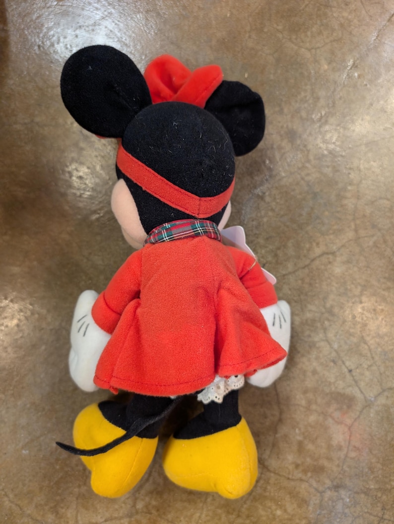 K&ouml;nnte beinhalten: Eine Pl&uuml;schpuppe von Minnie Mouse, von hinten gesehen, mit rotem Kleid, Schleife und Stirnband. Die Puppe hat schwarze Ohren, gelbe Schuhe, wei&szlig;e Handschuhe und einen karierten Kragen. Das Kleid hat eine wei&szlig;e Spitzentrim.