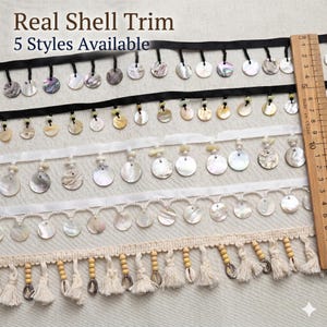 Op de afbeelding: Vijf stijlen schelpenband met ronde schelpschijfjes bevestigd aan linten of kralenstrengen. Een houten liniaal aan de rechterkant toont een breedte van ongeveer 10 centimeter. De tekst luidt Real Shell Trim 5 Styles Available.