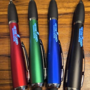 May include: Four retractable ballpoint pens in red, green, blue, and black with black rubber grips and stylus tips. Each pen features a silver pocket clip and the printed text BE THE LIGHT ILLUMINATE THE PAGE in white lettering on the barrel.