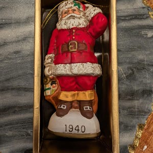 May include: A vintage Santa Claus Christmas ornament from 1940, presented in a gold box. The ceramic figure is dressed in a red suit with a white beard and fur trim. He carries a sack and is standing on a white base with the year 1940.