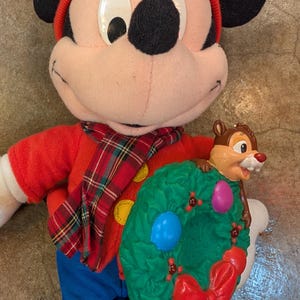 May include: A Mickey Mouse plush doll wearing a red hat, shirt, and plaid scarf, holding a green wreath with a red bow and a Chip figure. The doll has black ears and blue pants. The wreath has blue and pink ornaments.