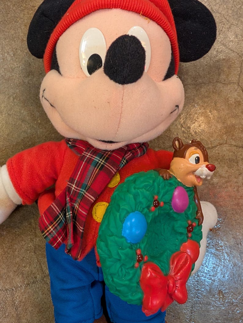 May include: A Mickey Mouse plush doll wearing a red hat, shirt, and plaid scarf, holding a green wreath with a red bow and a Chip figure. The doll has black ears and blue pants. The wreath has blue and pink ornaments.