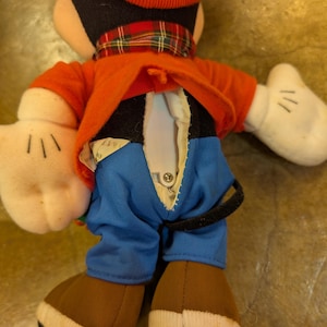 May include: A plush Mickey Mouse doll, viewed from the back, wearing a red hat and jacket, blue pants, and brown shoes. The doll has white gloves and a plaid scarf. The back of the doll's jacket is open, revealing a button closure.