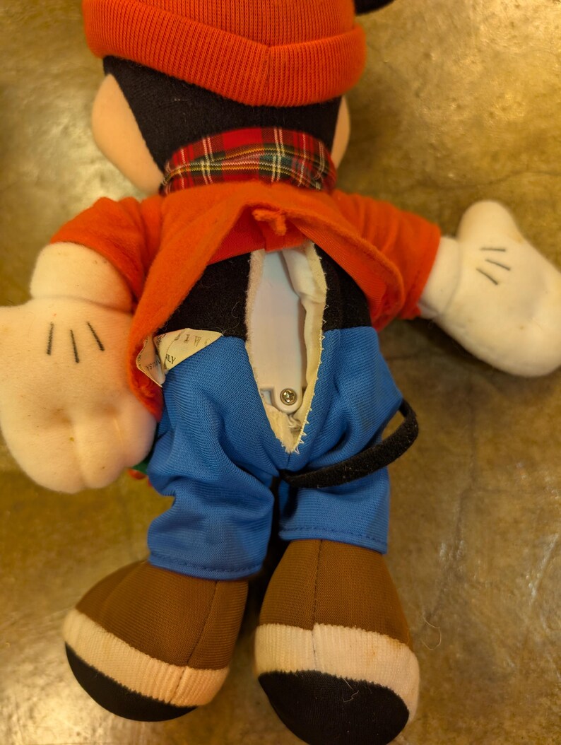 May include: A plush Mickey Mouse doll, viewed from the back, wearing a red hat and jacket, blue pants, and brown shoes. The doll has white gloves and a plaid scarf. The back of the doll's jacket is open, revealing a button closure.