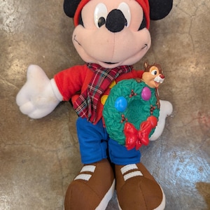 May include: A plush Mickey Mouse doll wearing a red hat, shirt, and scarf with a plaid pattern. The doll is holding a green wreath with colorful ornaments and a small Chipmunk figure. It is wearing blue pants and brown shoes.