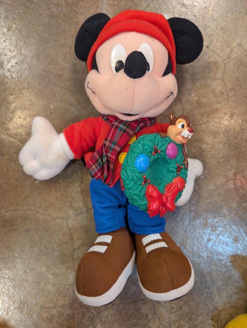 May include: A plush Mickey Mouse doll wearing a red hat, shirt, and scarf with a plaid pattern. The doll is holding a green wreath with colorful ornaments and a small Chipmunk figure. It is wearing blue pants and brown shoes.