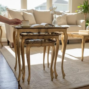 Puede incluir: Un juego de tres mesas nido doradas antiguas con patas curvas y ornamentadas. Las mesas están dispuestas frente a un sofá beige y una ventana. Una mano se extiende hacia una taza de té sobre la mesa superior. Mueble decorativo.