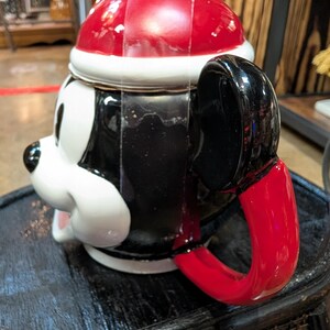 Puede incluir: Una taza de cer&aacute;mica con forma de cabeza de personaje de dibujos animados que lleva un gorro de Pap&aacute; Noel rojo y blanco. La taza presenta un acabado brillante en blanco y negro, un asa negra con forma de oreja y una base roja.