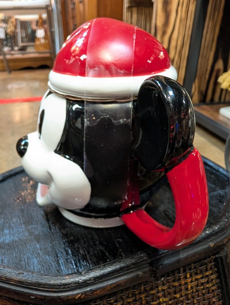 Puede incluir: Una taza de cer&aacute;mica con forma de cabeza de personaje de dibujos animados que lleva un gorro de Pap&aacute; Noel rojo y blanco. La taza presenta un acabado brillante en blanco y negro, un asa negra con forma de oreja y una base roja.