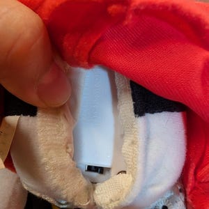 K&ouml;nnte beinhalten: Ein Pl&uuml;schtier mit rotem Hut und schwarz-wei&szlig;em K&ouml;rper. Das Spielzeug hat ein wei&szlig;es Batteriefach. Das Spielzeug wird ge&ouml;ffnet gehalten, um das Batteriefach zu zeigen.