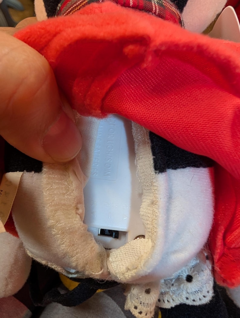 K&ouml;nnte beinhalten: Ein Pl&uuml;schtier mit rotem Hut und schwarz-wei&szlig;em K&ouml;rper. Das Spielzeug hat ein wei&szlig;es Batteriefach. Das Spielzeug wird ge&ouml;ffnet gehalten, um das Batteriefach zu zeigen.