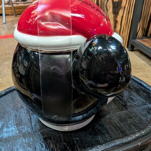 Puede incluir: Un tarro de galletas de cer&aacute;mica con forma de ping&uuml;ino que lleva un gorro de Pap&aacute; Noel rojo y blanco. El ping&uuml;ino tiene un cuerpo negro brillante y una barriga blanca. El texto Hand Painted es visible en el gorro rojo. Una correa de pl&aacute;stico transparente asegura la tapa a la base.