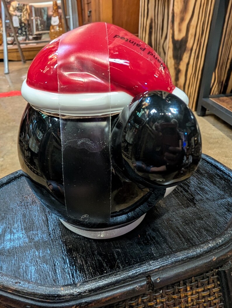Puede incluir: Un tarro de galletas de cer&aacute;mica con forma de ping&uuml;ino que lleva un gorro de Pap&aacute; Noel rojo y blanco. El ping&uuml;ino tiene un cuerpo negro brillante y una barriga blanca. El texto Hand Painted es visible en el gorro rojo. Una correa de pl&aacute;stico transparente asegura la tapa a la base.