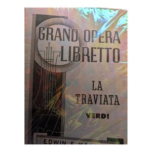 May include: A book cover for "Grand Opera Libretto" featuring the opera "La Traviata" by Verdi. The cover has a silver and iridescent sheen with black text and a stylized illustration of a stringed instrument.