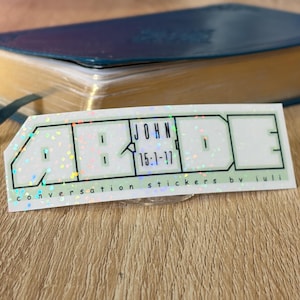 May include: A holographic sticker with the word "ABIDE" in a geometric font. The sticker also includes the text "JOHN 15:1-17" and "conversation stickers by iuli". The sticker is on a wooden surface.