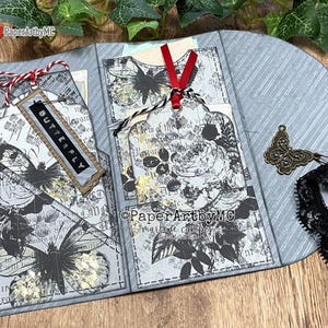 Może przedstawiać: Ręcznie wykonane szare rękodzieło z papieru z motywami motyli i kwiatów. Rękodzieło ma etykietę z napisem "BUTTERFLY". Jest ozdobione czerwoną i białą nitką, czarną koronkową wstążką i urokiem motyla.