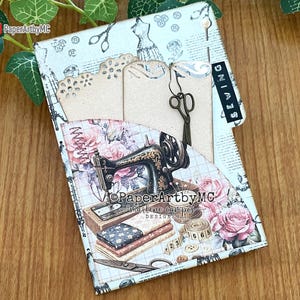 May include: A handmade sewing-themed journal with a vintage sewing machine illustration, roses, and fabric swatches. The journal includes tags, scissors, and the word "SEWING" on a tab. The background is a light blue and cream color.