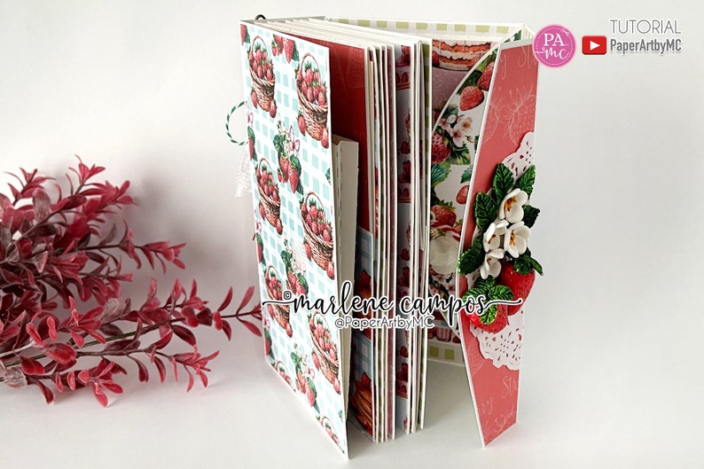May include: A handmade scrapbook album with a strawberry theme. The pages feature a repeating pattern of strawberry baskets on a blue and white checkered background. The album is decorated with paper strawberries and flowers.