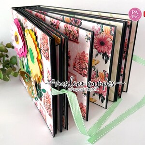 May include: A black scrapbook with a floral design and a green ribbon. The scrapbook has a 3D flower embellishment on the front. The text "marlene campos" is written on the front of the scrapbook.