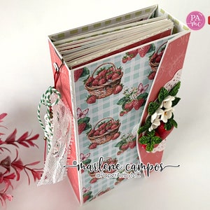 May include: A handmade paper craft album with a strawberry theme. The album features a light blue and white checkered pattern with strawberry illustrations. Embellishments include lace, twine, and paper flowers. The text "marlene campos" and "PaperArtybyMC" are visible.