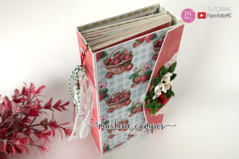 May include: A handmade paper craft album with a strawberry theme. The album features a light blue and white checkered pattern with strawberry illustrations. Embellishments include lace, twine, and paper flowers. The text "marlene campos" and "PaperArtybyMC" are visible.