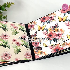 May include: A scrapbook with a floral design featuring pink and white flowers and butterflies. The scrapbook is open to reveal a page with a white background and a floral design featuring pink, yellow, and orange flowers and butterflies. The text "marlene campos" and "@PaperArtbyMC" is printed on the page.